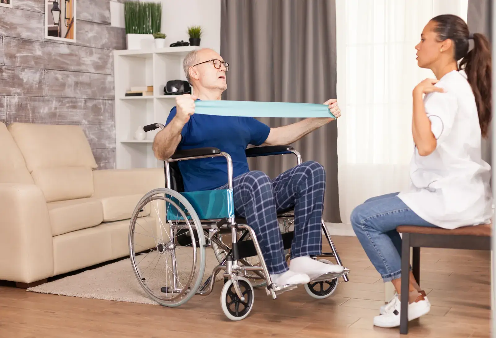 Älterer Mann im Rollstuhl macht zu Hause mit einer Pflegekraft Kräftigungsübungen mit einem Übungsband