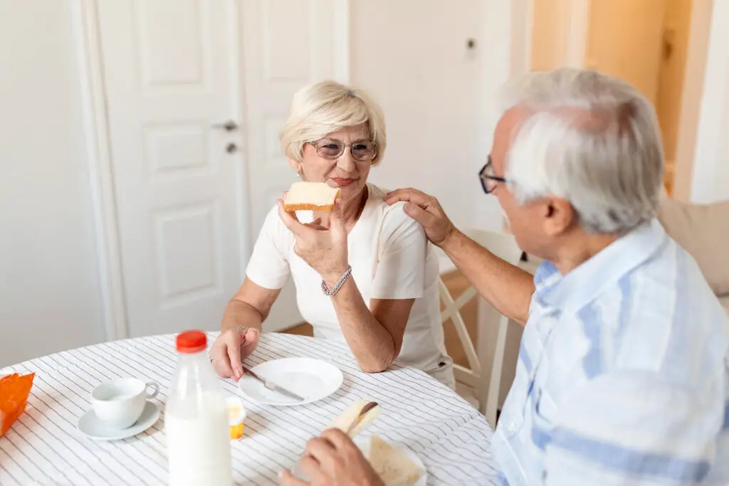 Seniorenpaar sitzt zuhause am Frühstückstisch, die Frau isst ein Stück Brot