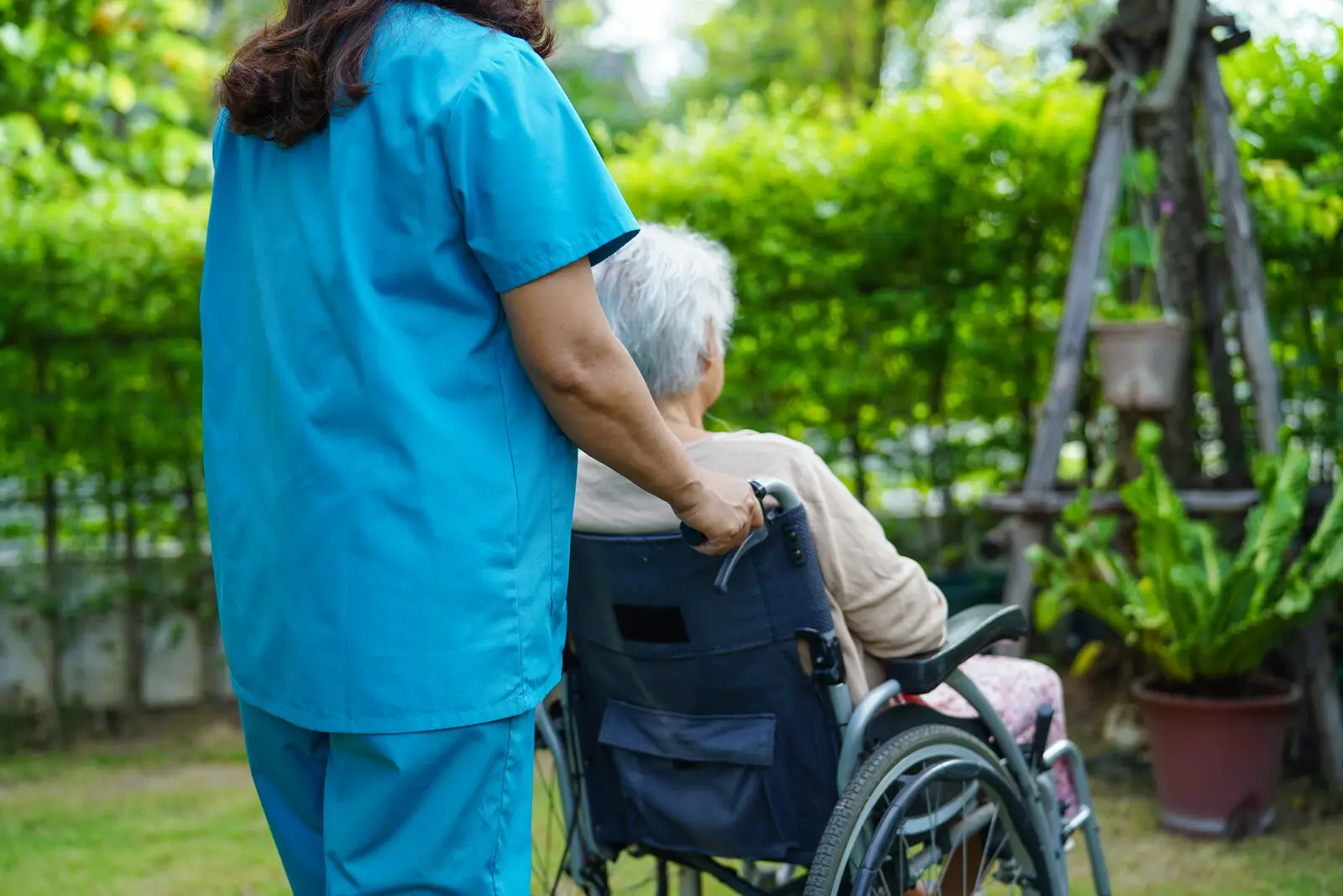 Pflegekraft begleitet Seniorin im Rollstuhl im Park, Betreuung nach bewilligtem Pflegegrad