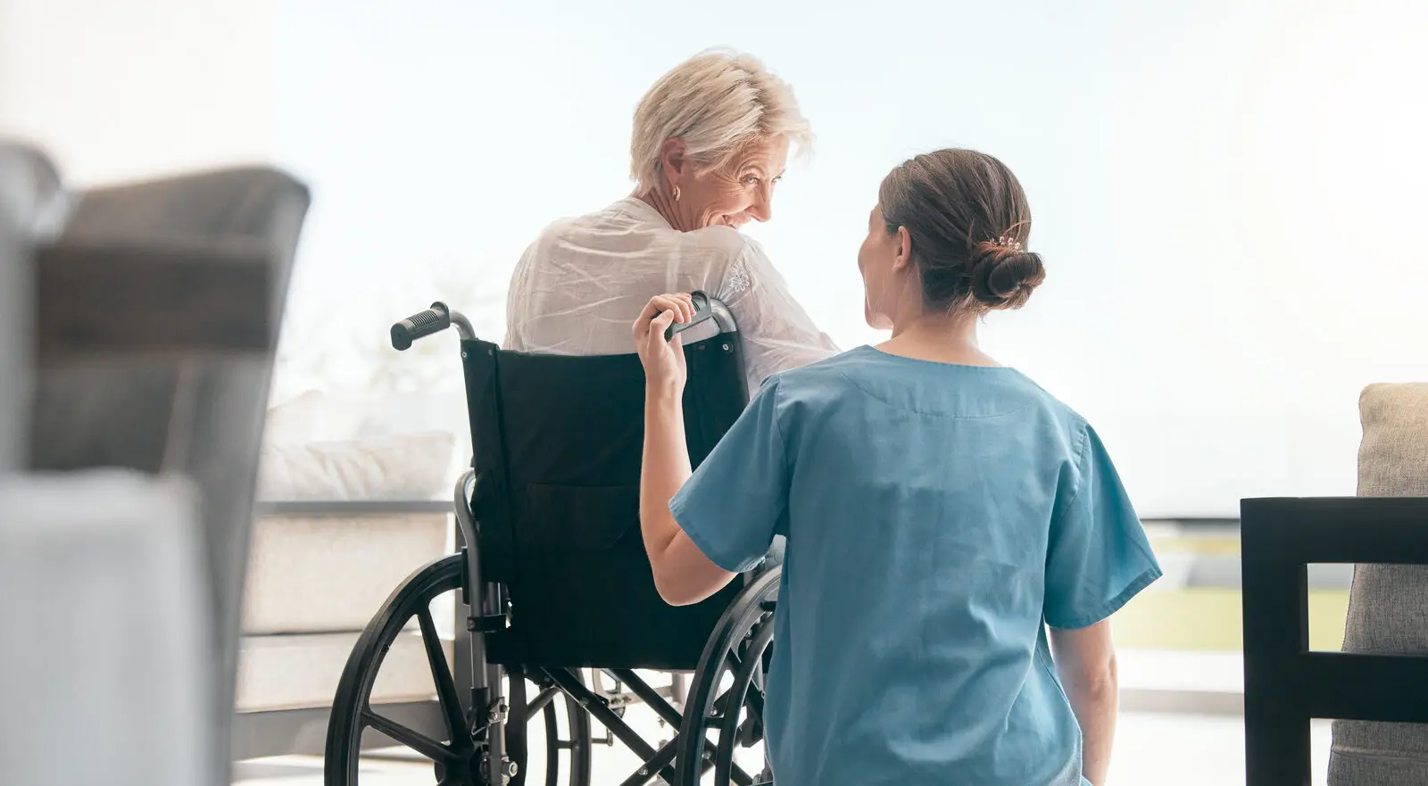 Pflegekraft im Gespräch mit lächelnder Seniorin im Rollstuhl über Schmerzen im Alter